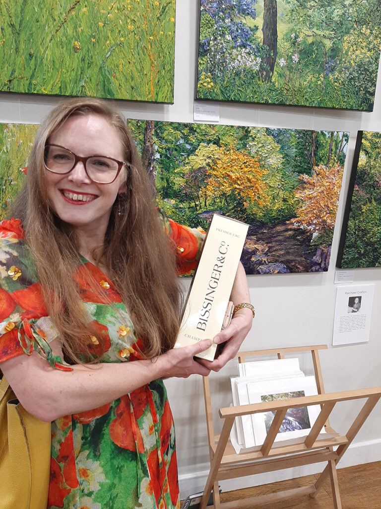 Artist smiling in front of a display of their art - holding an award received for best work in the show