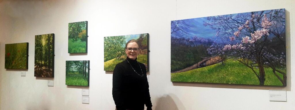 The artist stands before her paintings in an exhibition space & smiles