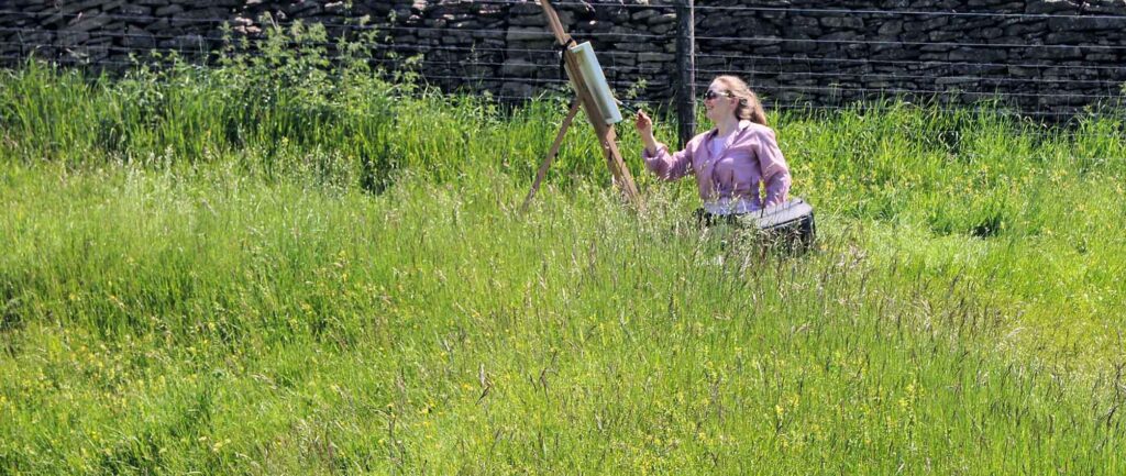 Artist appears with easel and canvas in the distance in a sunny field